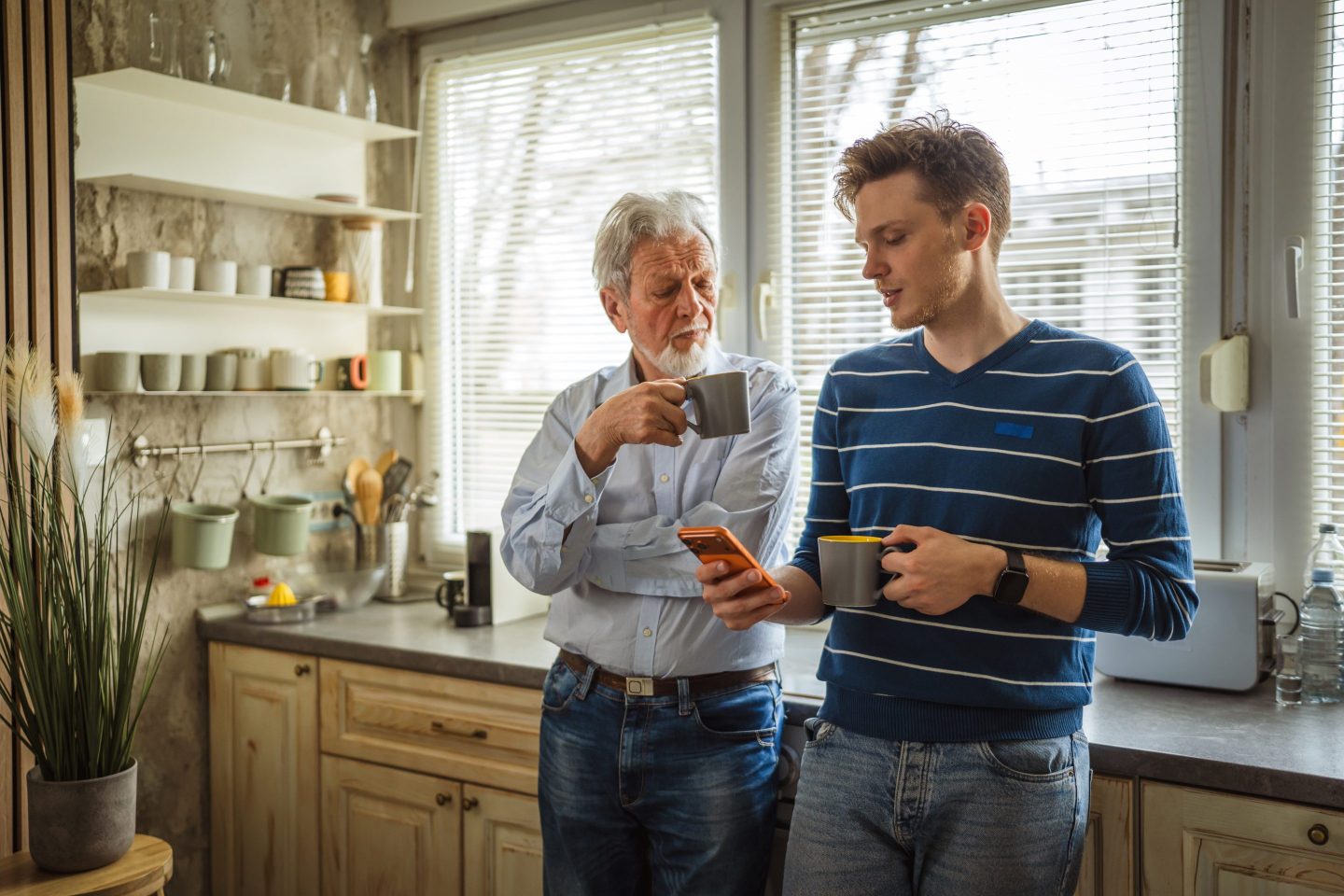 A young man holding a cup of coffee in a kitchen shows his phone to an older man, who is also holding a mug.