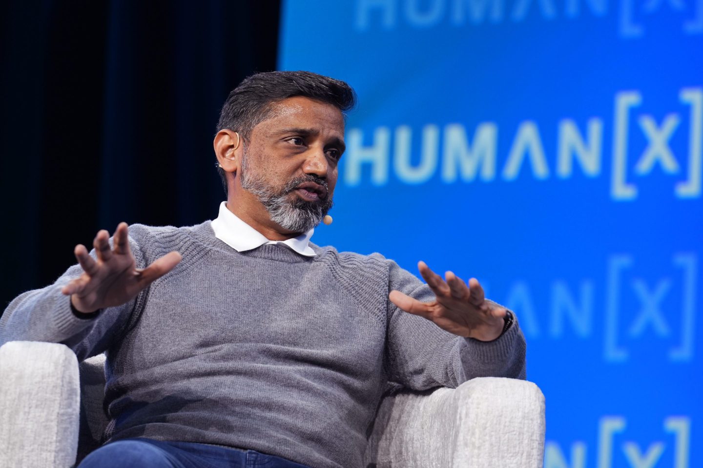 Praveen Neppalli Naga sits in a white chair in front of a blue background onstage.